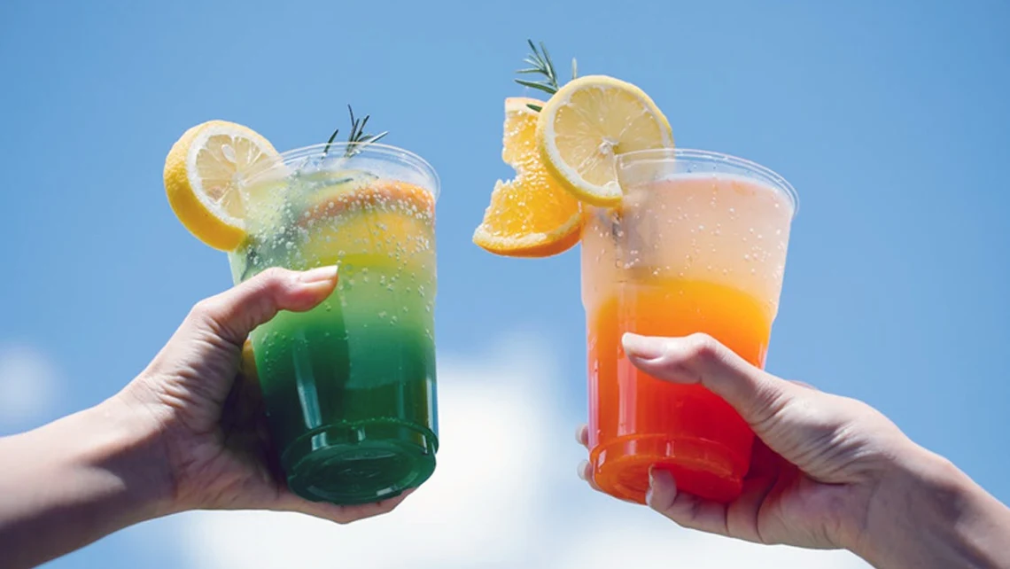 Hands holding lemon-garnished green and orange adaptogen drinks against a bright blue and white sky