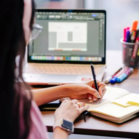 A girl writing in her notebook while facing an open laptop beside a jar of pens and highlighters