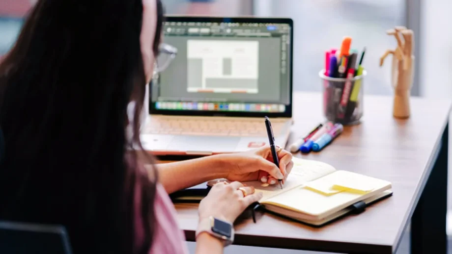 A girl writing in her notebook while facing an open laptop beside a jar of pens and highlighters