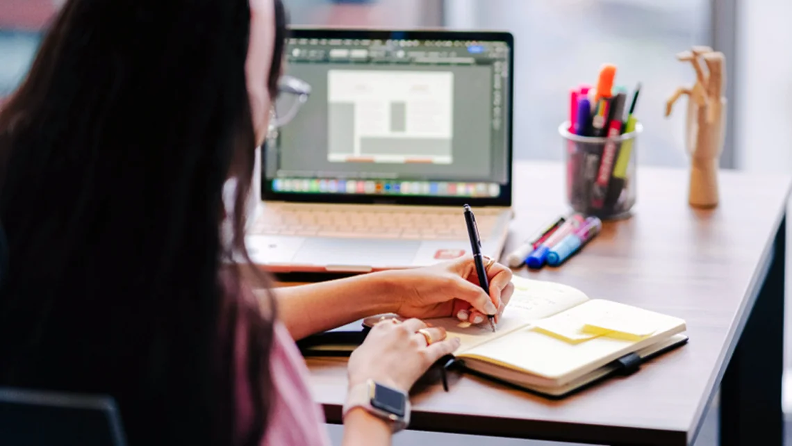 A girl writing in her notebook while facing an open laptop beside a jar of pens and highlighters
