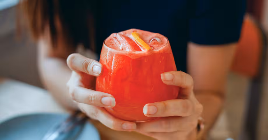 close-up shot of a woman’s hands holding a glass filled to the brim with a red-orange THC drink