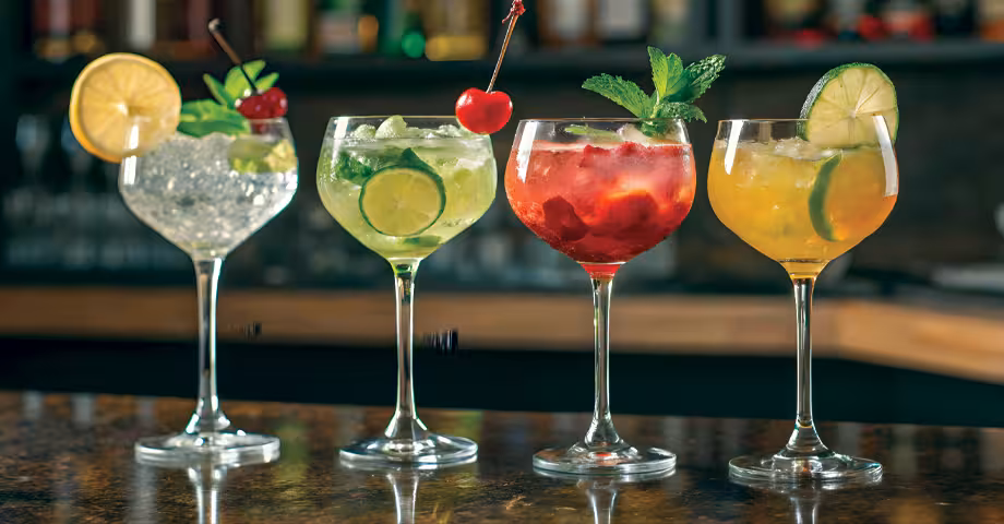 four wine glasses of mocktails with fruit and herb garnishes lined on a bar counter