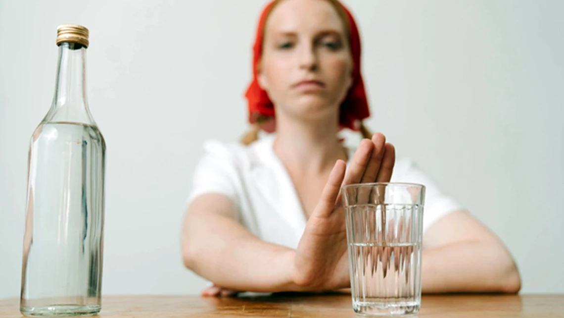 woman in red bonnet pushing away a glass of alcohol beside a liquor bottle on a table