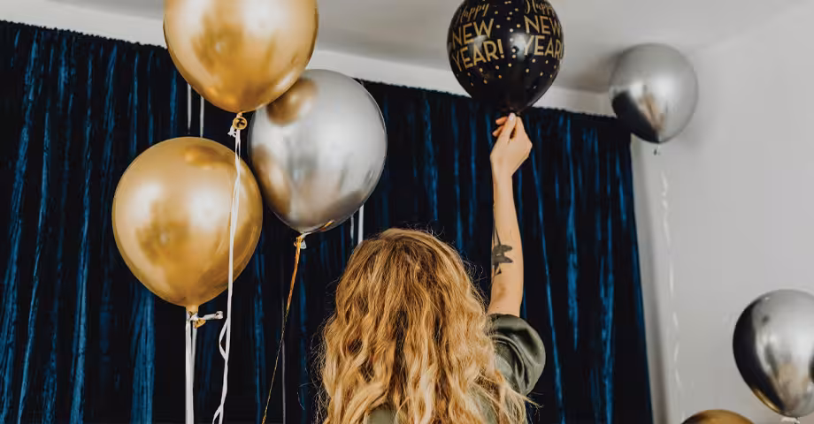 woman sticking a “happy new year” balloon to the ceiling along with other gold and silver balloons