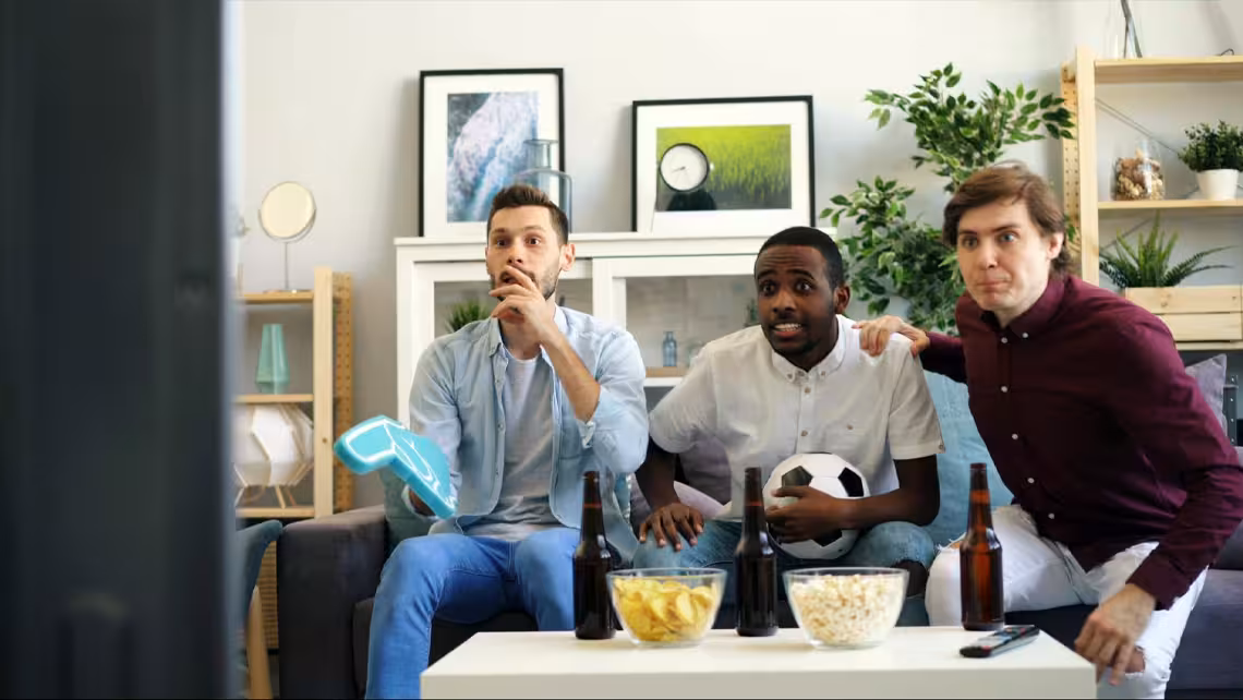 3 guys on a couch watching TV with the best Super Bowl drinks and snacks on a table