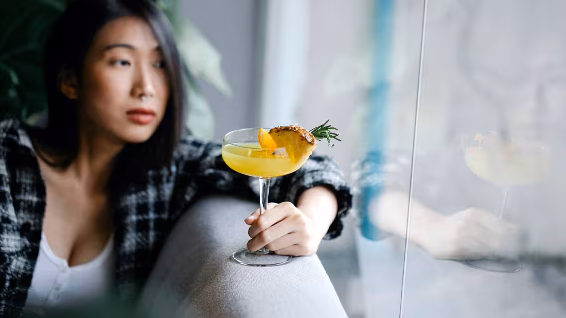 Asian woman leaning on a couch looking outside a window holding a pineapple mocktail